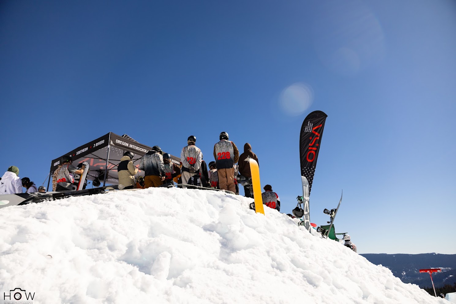 Photo of top of of course at a halfpipe contest with snowboarders standing around. The foreground is snow, background blue sky.
